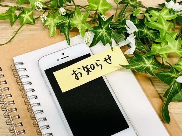 机の上にあるノートとスマホに貼られたお知らせの付箋